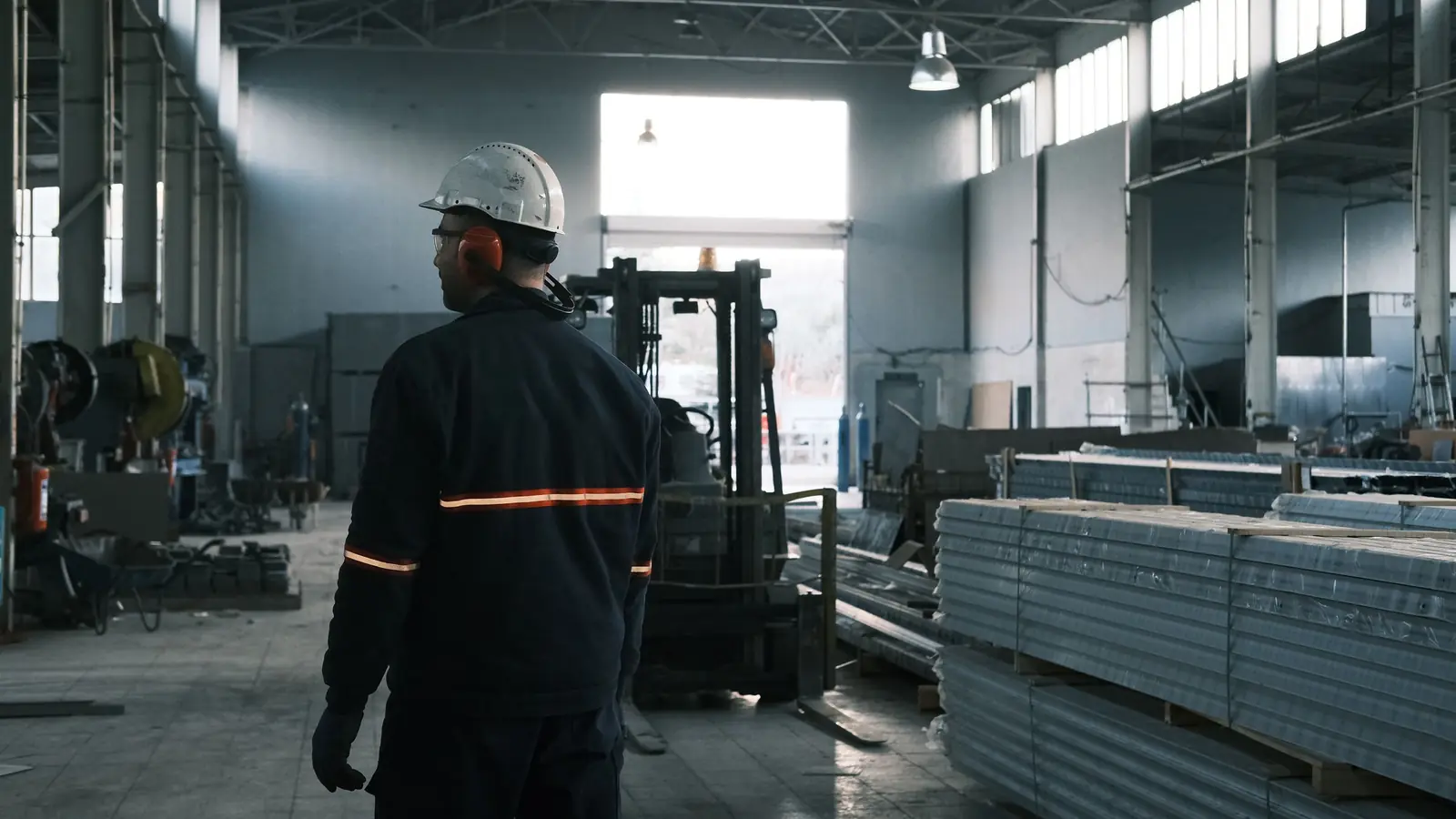 Mitarbeiter mit Schutzhelm in einer Werkshalle mit Stapler und Materiallager